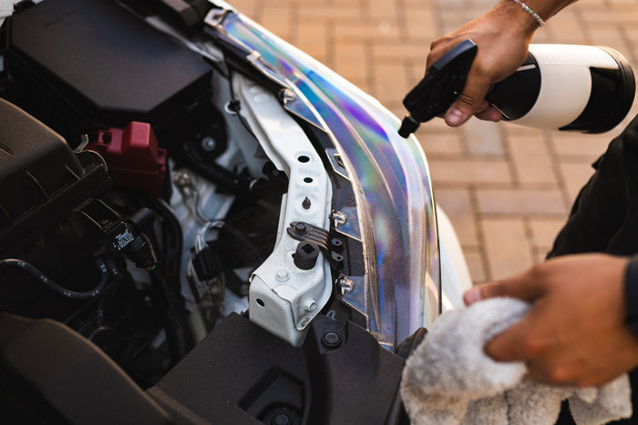 A person is cleaning the edge of a car's engine bay using a spray bottle filled with Koch Chemie Wash & Finish and a microfiber towel, with focus on the metallic frame and iridescent finish near the hood area.