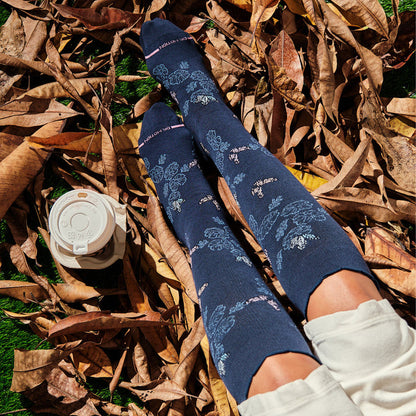 Knee-high compression socks with a navy background and whimsical butterfly and floral designs, resting on dry leaves.